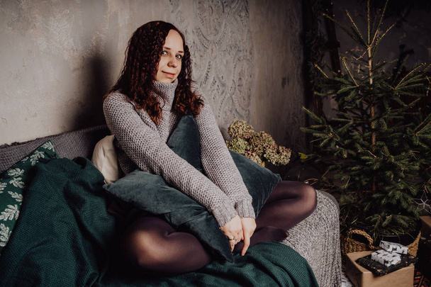 A young girl in a sweater sitting on the bed huddled in a blanket. Woman dressed in warm sweater, street style clothing. Clean skin, long hair - Zdjęcie, obraz