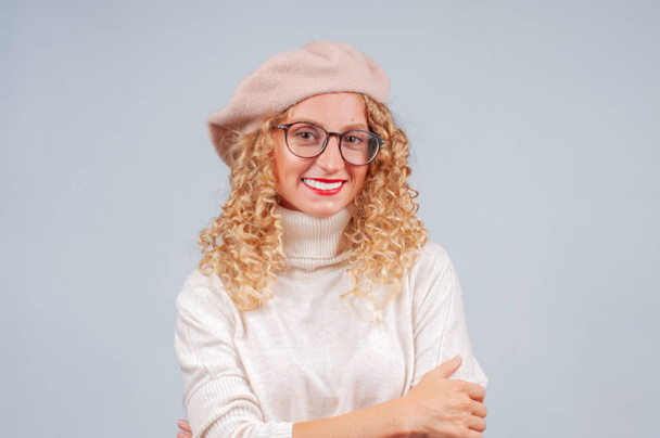 Portrait of a student woman with curly hair dressed in warm sweater, beret and eyeglasses looking at camera on gray background - Foto, afbeelding