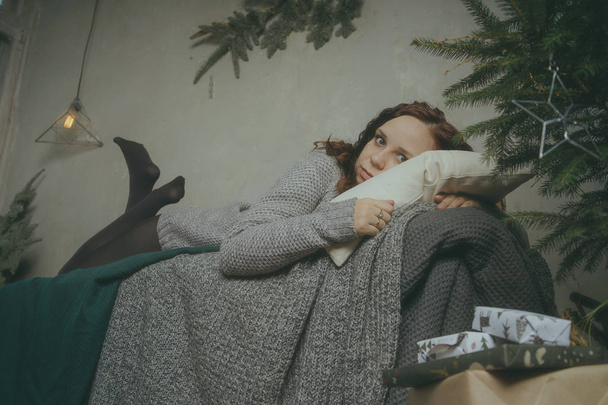 Winter Concept. Cute young girl in a gray knitted sweater. Beautiful woman is relaxing in a white bedroom. Beautiful women in clothes are waking up in the morning. Woman wearing a sweater - Foto, Imagen