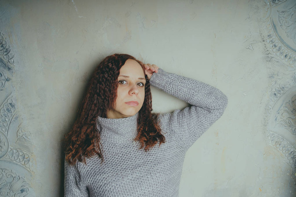 A young girl in a sweater posing against a gray wall. Woman dressed in warm sweater, street style clothing. Clean skin, long hair - Фото, зображення