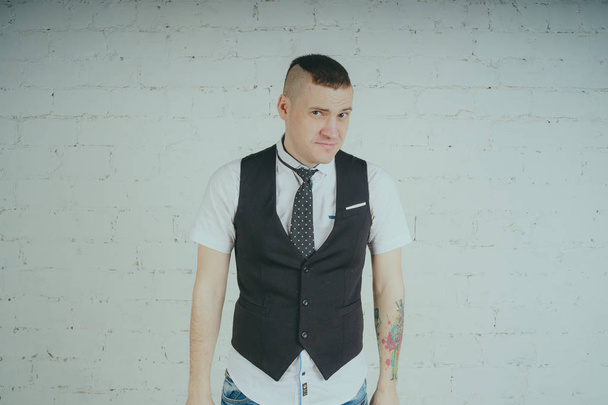 Emotional office worker with a tattoo, posing against a white brick wall. The man is dressed in jeans and a white shirt with a tie. - Foto, Imagen