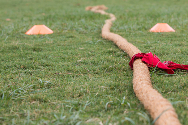 Corda grossa com bandana vermelha anexada encontra-se na grama antes do início do concurso de cabo de guerra
 - Foto, Imagem
