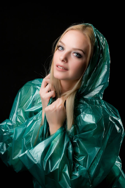Portrait of a young blond woman in a green raincoat looking at the camera, a hood is put on her head. Shooting in a photo studio on a black background - Photo, Image