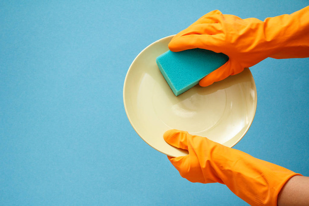 Women's hands in orange protective gloves with plate and sponge on blue background. Washing and cleaning concept. - 写真・画像