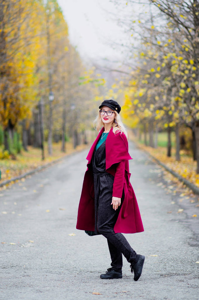 Linda mujer joven en gafas y gorra, con un abrigo rojo caminando en el parque en el otoño del año
. - Foto, Imagen