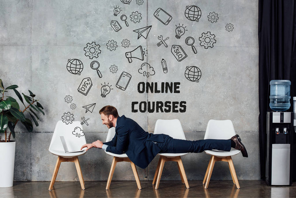 businessman lying on chairs and using laptop in waiting hall with online courses lettering on wall - Photo, Image