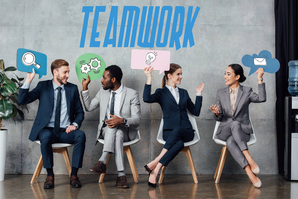 happy multiethnic businesspeople holding speech bubbles with teamwork icons while sitting in waiting hall - Foto, immagini