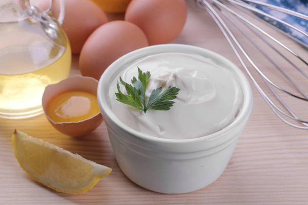 Mayonnaise in a white ceramic bowl and ingredients for making mayonnaise on a natural wooden background. white sauce. - Photo, Image