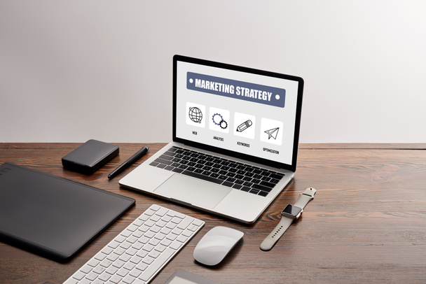 flache Lage von Gadgets und Laptop mit Marketing-Strategie-Website auf dem Bildschirm am Arbeitsplatz isoliert auf grau - Foto, Bild