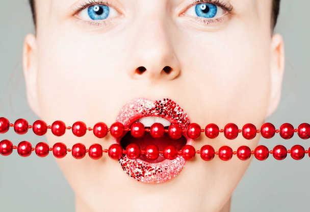 Kreatives Porträt eines Frauengesichts mit leuchtenden Lippen und roten Perlen in Nahaufnahme. perfekter weiblicher Mund mit glitzerndem Lippenstift Make-up  - Foto, Bild
