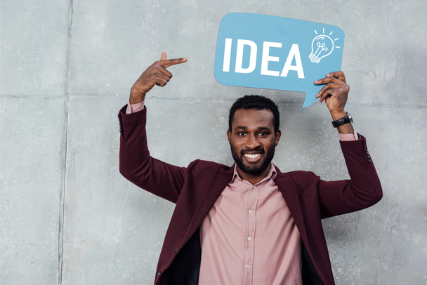smiling african american casual businessman looking at camera and pointing with finger at speech bubble with idea lettering and light bulb icon - Photo, Image