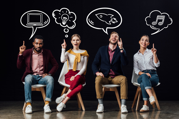 multiethnic group of people sitting on chairs and showing idea gestures with speech and thought bubbles above heads isolated on black - Photo, Image