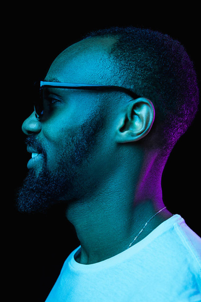 Close up portrait of a young happy smiling african man in sunglasses at studio. High Fashion male model in colorful bright lights posing on black background. Art design concept - Foto, Imagem