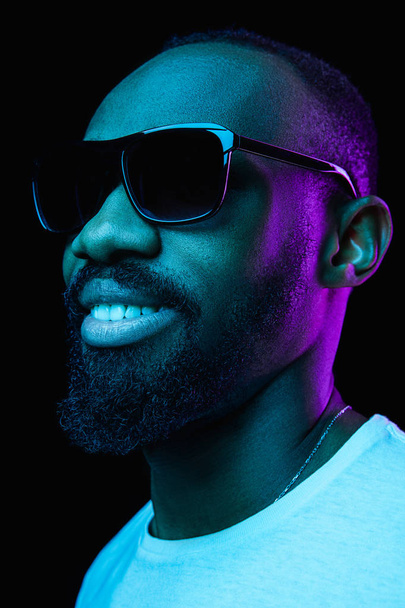 Close up portrait of a young happy smiling african man in sunglasses at studio. High Fashion male model in colorful bright lights posing on black background. Art design concept - Photo, Image