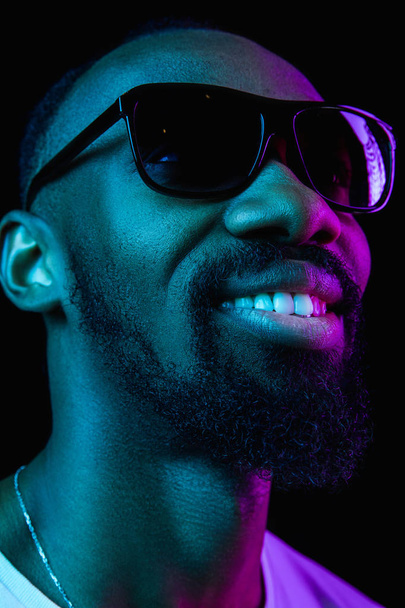 Close up portrait of a young happy smiling african man in sunglasses at studio. High Fashion male model in colorful bright lights posing on black background. Art design concept - Foto, Imagen