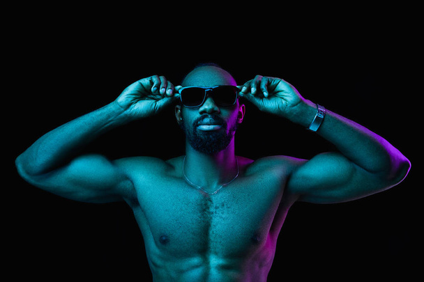 Close up portrait of a young naked happy smiling african man in sunglasses at studio. High Fashion male model in colorful bright lights posing on black background. Art design concept - Photo, Image
