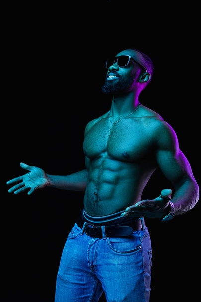 Close up portrait of a young naked happy smiling african man in sunglasses at studio. High Fashion male model in colorful bright lights posing on black background. Art design concept - Zdjęcie, obraz