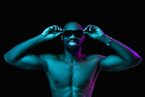 Close up portrait of a young naked happy smiling african man in sunglasses at studio. High Fashion male model in colorful bright lights posing on black background. Art design concept - Photo, image
