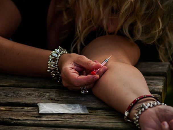 Uma mulher injetando droga em seu braço com uma seringa na rua e uma dose de droga para o lado
 - Foto, Imagem