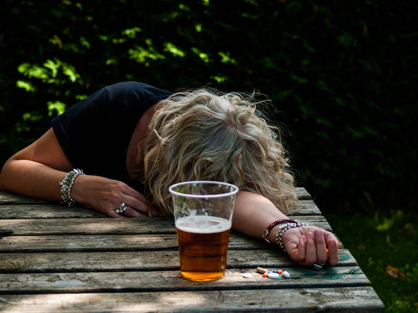 Uma mulher com um grande número de comprimidos na mão de diferentes cores e tamanhos e um copo de cerveja, em uma mesa ao ar livre
 - Foto, Imagem
