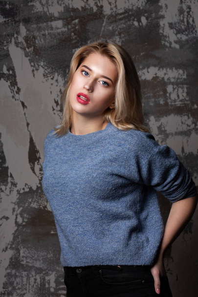 Stunning blonde woman with lush hair wearing blue sweater, posing in studio with shadows - Photo, Image