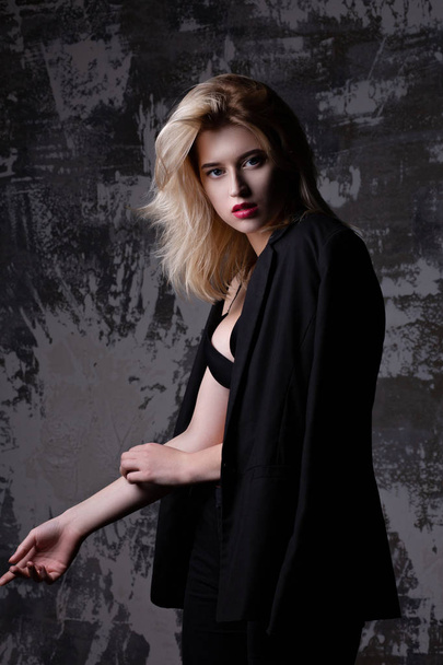 Gorgeous blonde model with perfect makeup wearing black jacket and bra posing at studio with shadows - Photo, Image