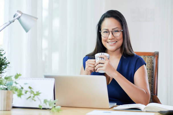Adult smiling Asian woman in glasses holding cup of coffee sitting at table with laptop in modern office smiling at camera - 写真・画像
