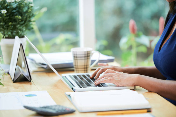 Vista lateral da colheita da mulher na mesa no escritório moderno usando laptop à luz do dia
  - Foto, Imagem
