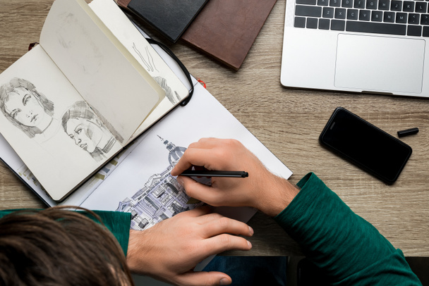 overhead back view of man drawing in album on wooden table next to laptop and smartphone - Photo, Image