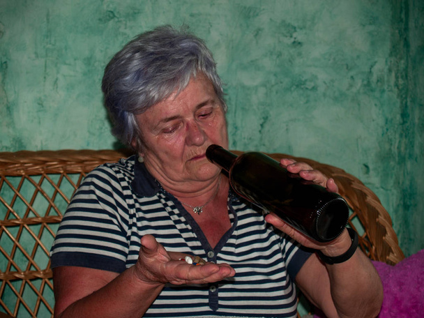 Een senior vrouw met een fles wijn en een groot aantal pillen in haar hand van verschillende kleuren en maten - Foto, afbeelding