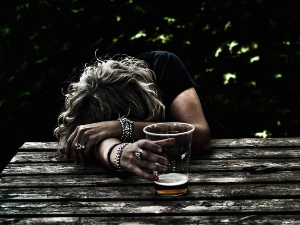 Una mujer borracha con un vaso de cerveza en la mano, inconsciente, apoyada en una mesa
 - Foto, Imagen