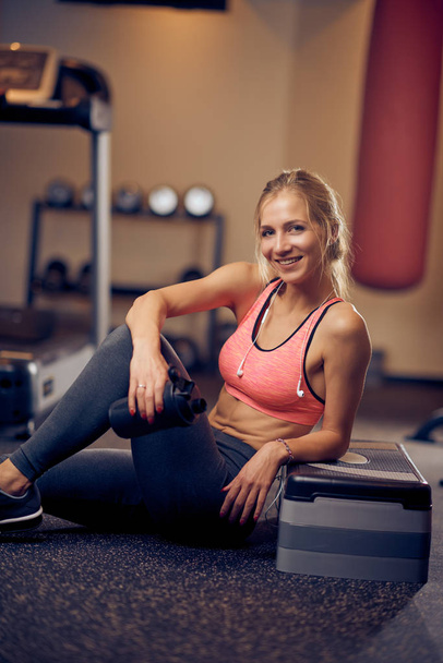 Mujer sonriente bebiendo agua y descansando después de hacer ejercicios mientras está sentada en el suelo. Interior del gimnasio
. - Foto, imagen