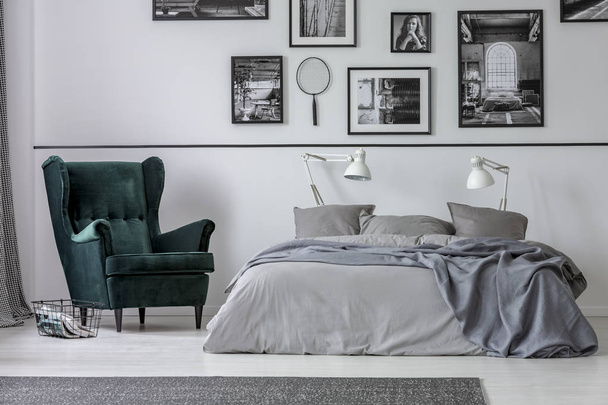 Armchair next to grey bed with white lamps in bedroom interior with gallery on the wall. Real photo - Foto, Imagem