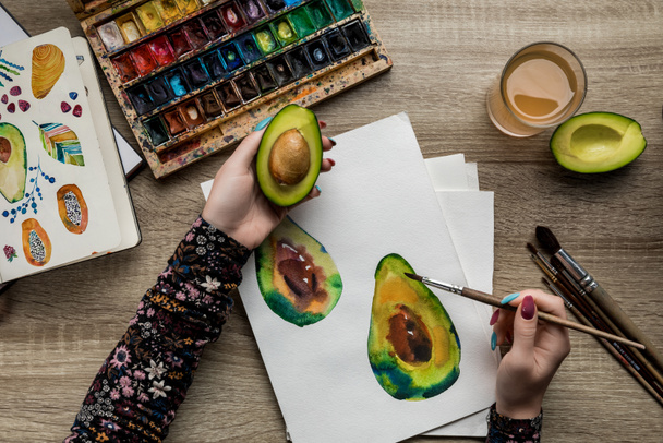 top view of female hands drawing avocado with watercolor paints and paintbrush on wooden table - Foto, Bild