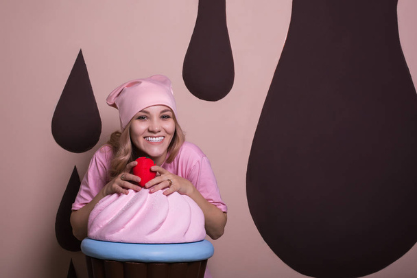 entzückendes blondes Mädchen mit pinkfarbener Mütze genießt große Cupcake im Studio. Raum für Text - Foto, Bild