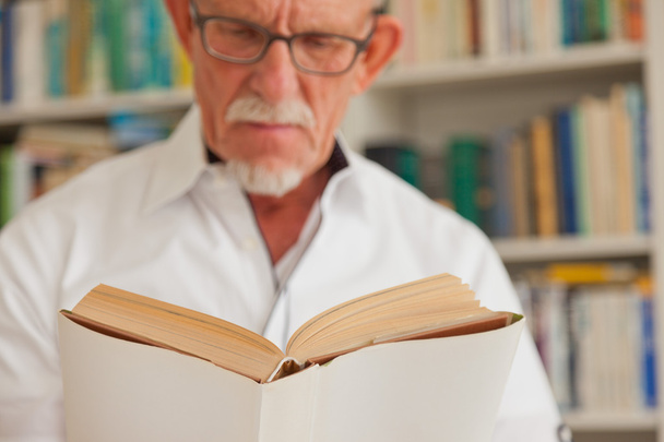 Senior mit Brille liest Buch vor Bücherregal. - Foto, Bild