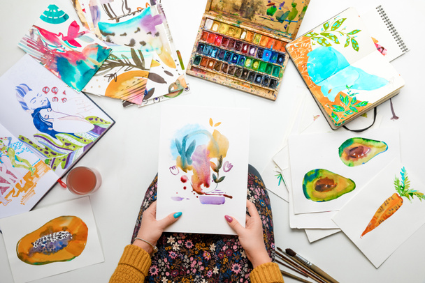 top view of woman holding watercolor drawing while surrounded by color pictures - Photo, Image