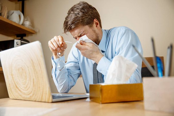 Sick man with handkerchief sneezing blowing nose while working in office, businessman caught cold, seasonal flu. Pandemic influenza, disease prevention, air conditioning in office cause sickness - Foto, imagen