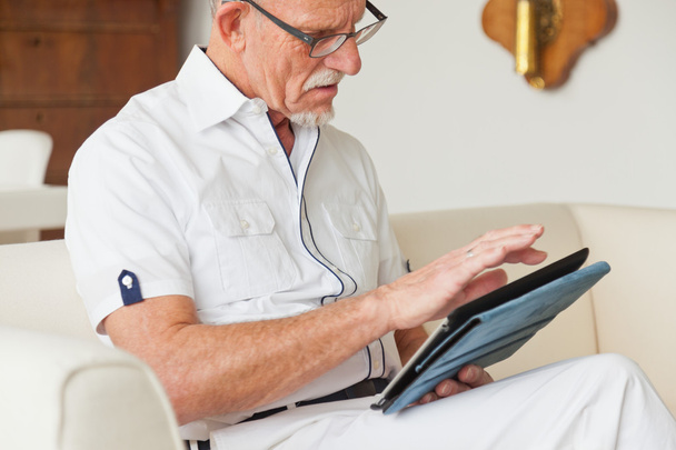 Homme âgé avec des lunettes en utilisant une tablette sur le canapé dans le salon
. - Photo, image