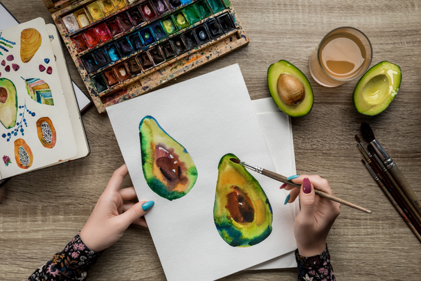top view of female hands drawing avocados with watercolor paints and paintbrush on wooden table - Photo, Image