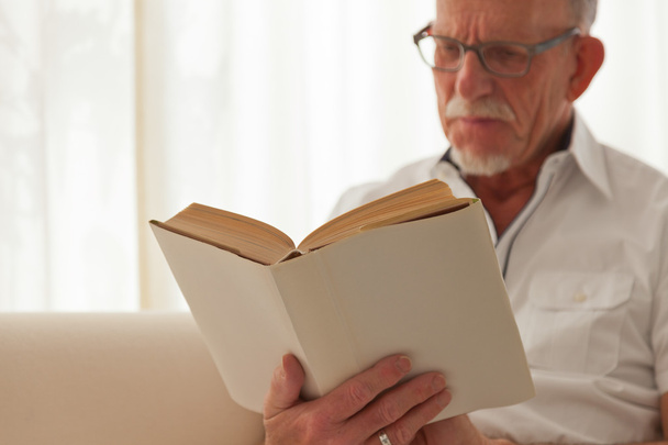 starszy człowiek w okularach, czytanie książki w salonie. - Zdjęcie, obraz