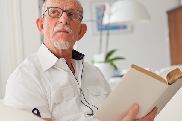 Senior man with glasses reading book in living room. - Foto, Bild