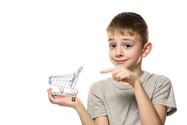 Aranyos fiú rendelkezik kis fém bevásárlókocsi a kezében, és rámutat arra, mutatóujjával. Izolálja a fehér háttér. Vásárlási koncepció - Fotó, kép
