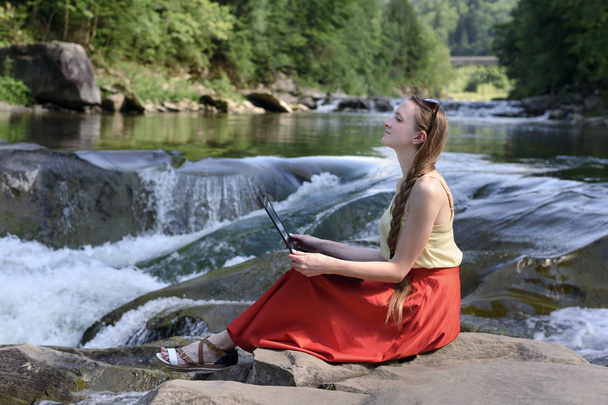 Ονειρεύεται το όμορφο μακρυμάλλης κορίτσι σε μια κόκκινη φούστα κάθεται με με ένα φορητό υπολογιστή σε μια πέτρα με φόντο ένα χείμαρρο από ένα ποτάμι βουνό. Ανεξάρτητος έννοια. Εργασία στη φύση. - Φωτογραφία, εικόνα