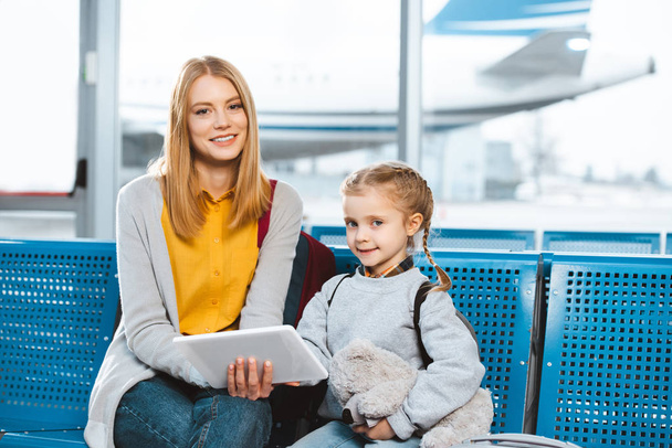 mooie moeder bedrijf digitale tablet en zitten in de luchthaven met dochter  - Foto, afbeelding