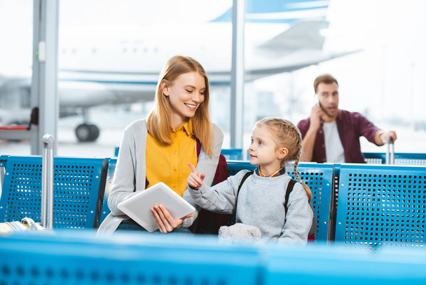 enfoque selectivo de la madre sosteniendo la tableta digital y sonriendo mientras mira a la hija en el aeropuerto
 - Foto, imagen