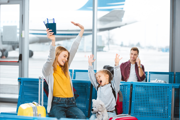 enfoque selectivo feliz madre e hija tomados de la mano por encima de la cabeza y sonriendo en el aeropuerto con el hombre en el fondo
 - Foto, imagen