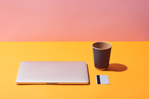 Kaffeetasse aus Papier in der Nähe von Laptop und Kreditkarte auf gelber Oberfläche und rosa Hintergrund - Foto, Bild