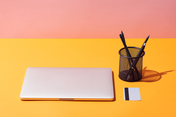  Bleistifthalter in der Nähe von Laptop und Kreditkarte auf gelber Oberfläche und rosa Hintergrund - Foto, Bild