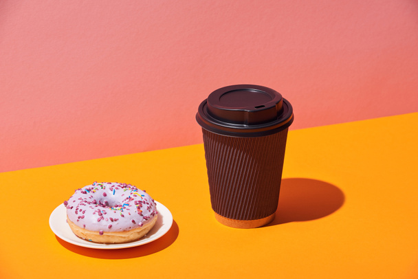 tasty donut with saucer and paper coffee cup on yellow surface and pink background - Фото, изображение